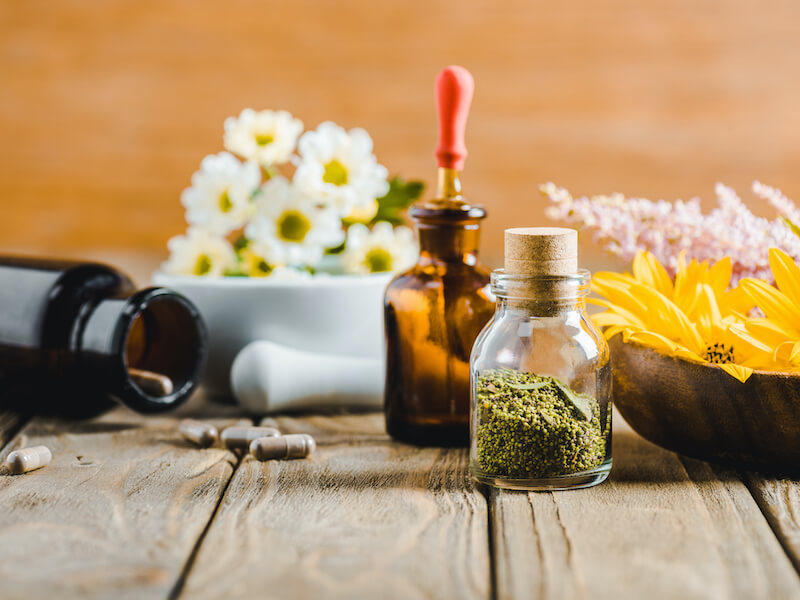 Homeopathic herbs and flowers in mortar and pestle with tincture and medicine bottles on a wooden table.