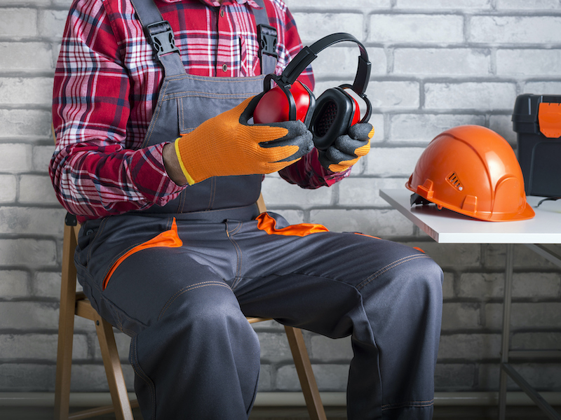 Worker sitting on a folding chair wearing a red plaid shirt and work overalls getting ready to put protective headphones on.