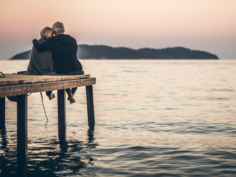 Older couple cuddling while sitting at the end of a dock overlooking a lake watching the sun set above mountains in the distance.