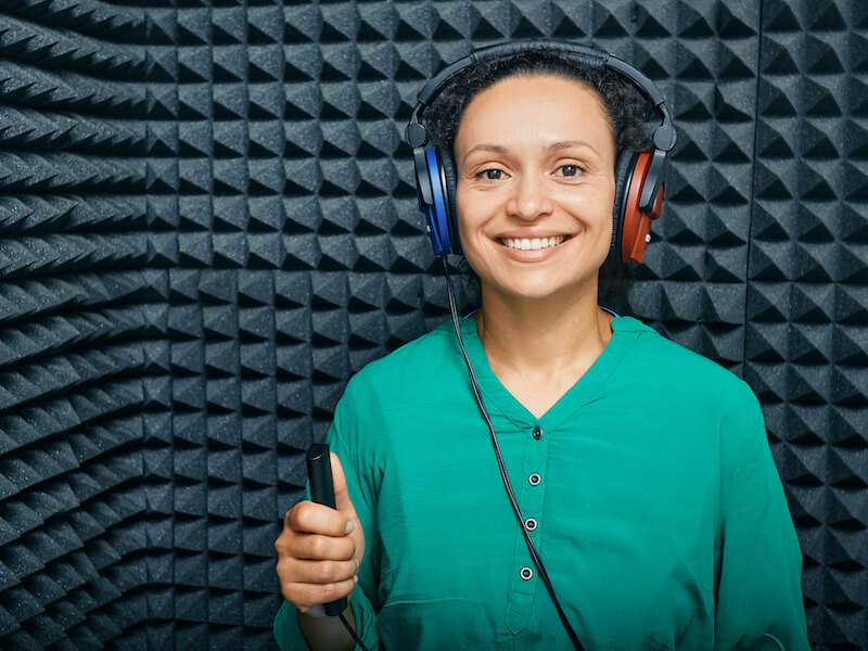 Smiling woman with short curly black hair wearing a green button up shirt excitedly waiting for her hearing test to begin in a sound booth