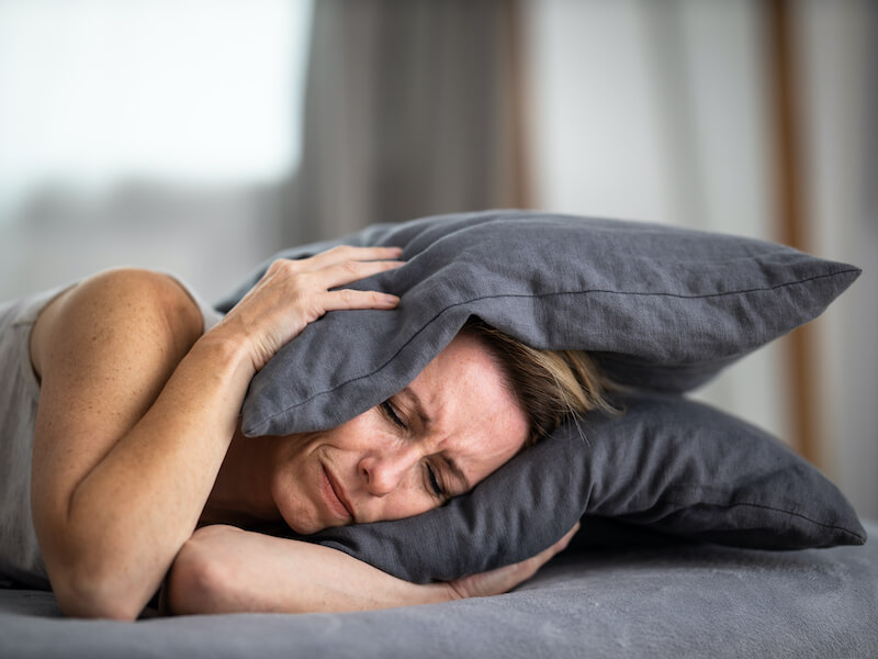 Woman suffering with tinnitus and grimacing laying down in bed pressing a gray pillow to her ears.