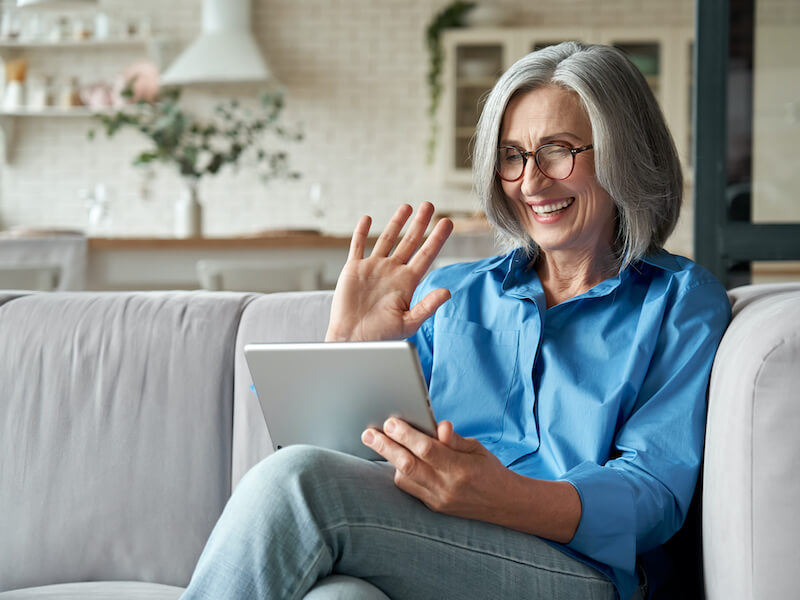 Happy mature middle aged adult woman wearing hearing aids waving hand holding digital tablet computer video conference calling by social distance virtual family online chat meeting sitting on couch at home