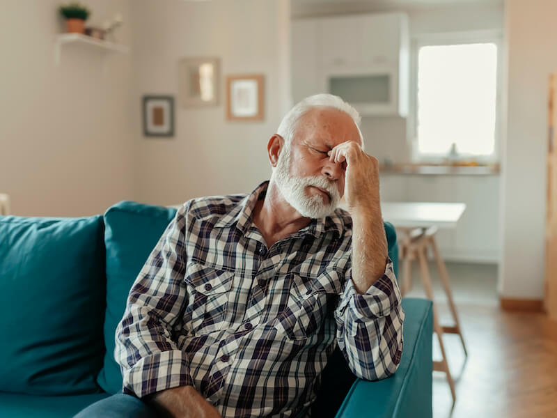 A distraught senior man sitting on his couch suffering from a headache because his hearing aids were not properly adjusted.