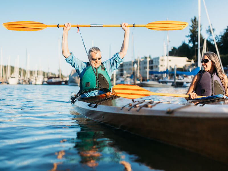 Older man in kayak in the harbour raising his paddle in the air in youthful excitement because he treated his hearing loss and feels youthful.