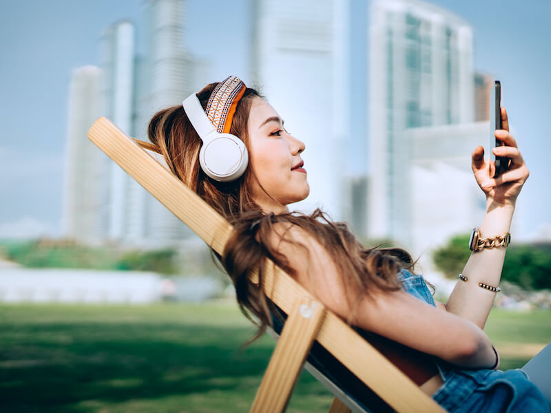 Woman with long dark hair relaxing in a chair in the park listening to headphones