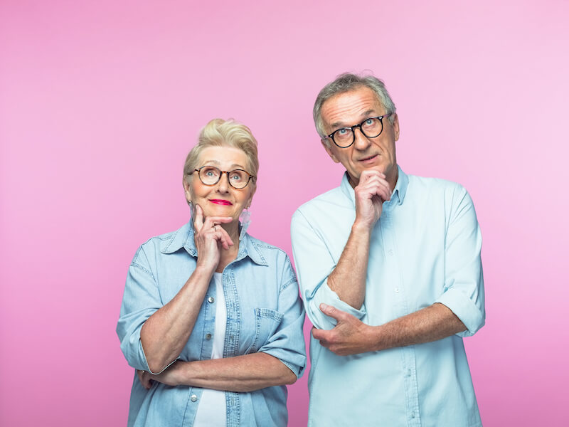 Senior couple suffering from hearing loss standing in front of a pink backdrop trying to remember something.
