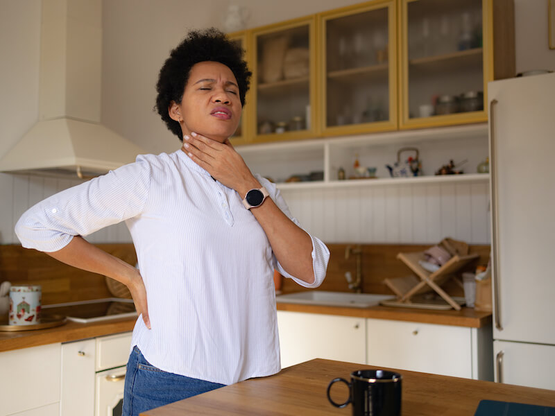 Woman feeling discomfort in her throat from laryngitis while standing in her kitchen.