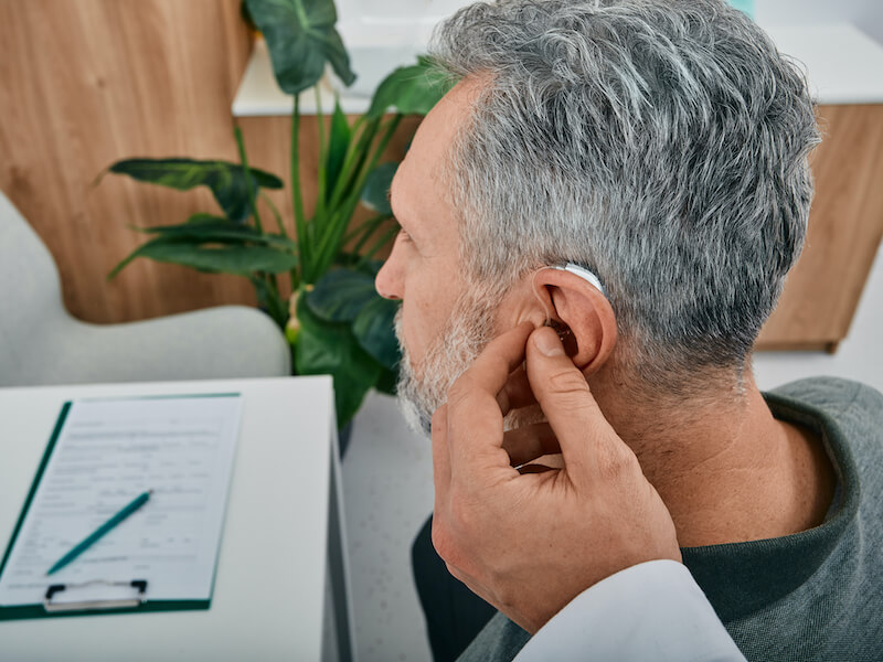 Audiologist fits a hearing aid on mature man ear while visit a hearing clinic.