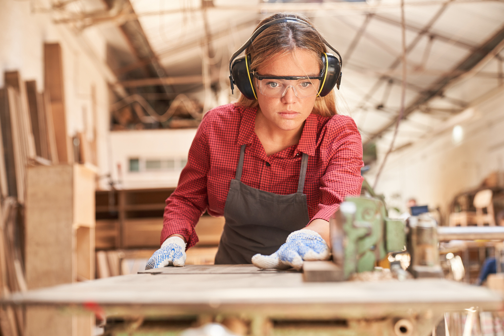 Young woman as a carpenter with safety glasses and hearing protection works on machine.